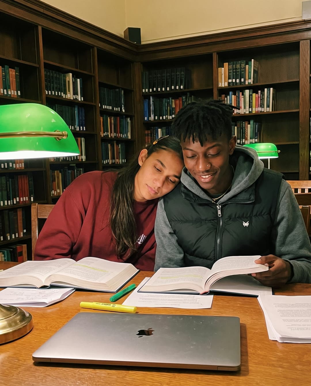 Ren resting her head on Kavi's shoulder as they study at a library table under a green lamp.