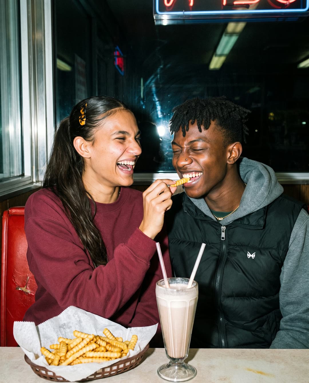Ren feeding Kavi a fry in a diner booth beside a milkshake, both laughing.